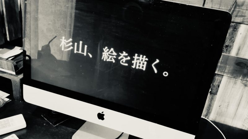 パソコンに、“杉山、絵を描く。”という文字が写っているモノトーンの写真