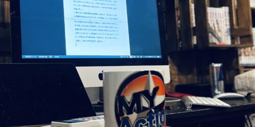 ”My Action”のロゴがプリントされているマグカップの奥に、パソコンのモニターに写っている書きかけの小説