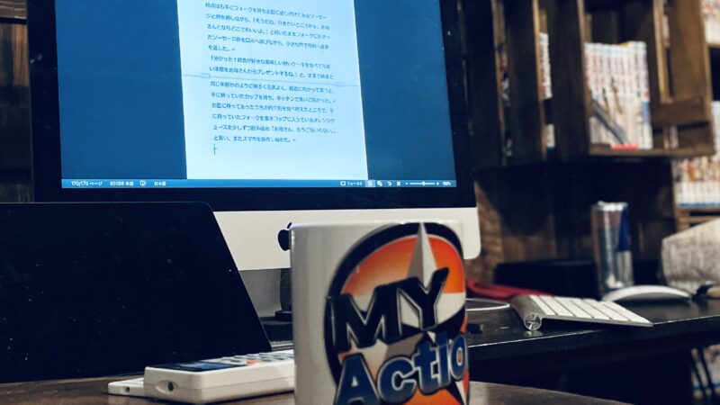 ”My Action”のロゴがプリントされているマグカップの奥に、パソコンのモニターに写っている書きかけの小説