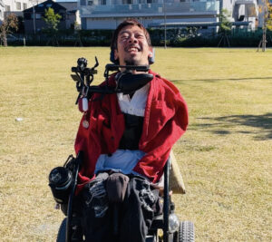 赤いジャケットを肩に羽織ってダメージジーンズ姿で電動車椅子に乗って、芝生の上にいる僕