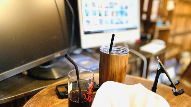 コーヒーが入ったコップとタンブラーの奥にパソコンが置いてある写真